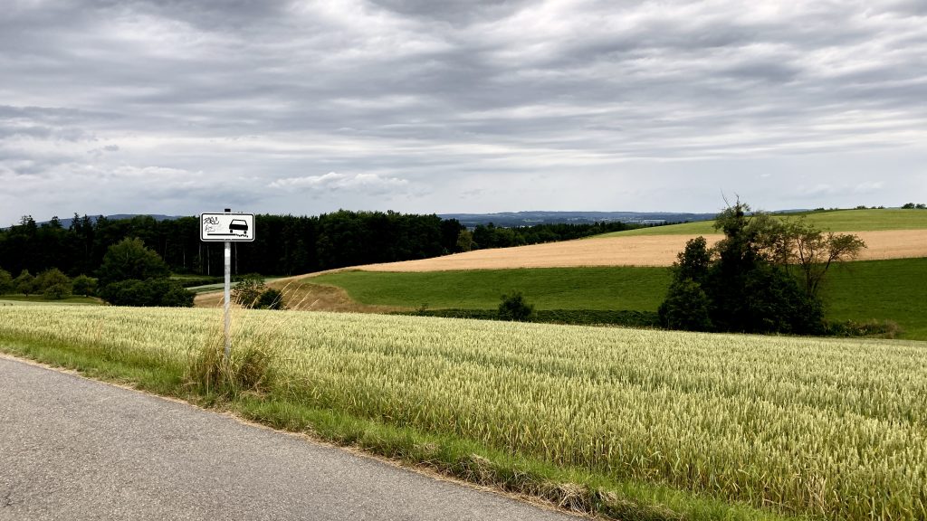 Image from July 2021 depicting the fields around Ravensburg in Southern Germany. There is a sign with some graffiti on which depicts a car sliding off the road, presumably because there is very little curb and it's likely you will career into the grass if you're not careful.